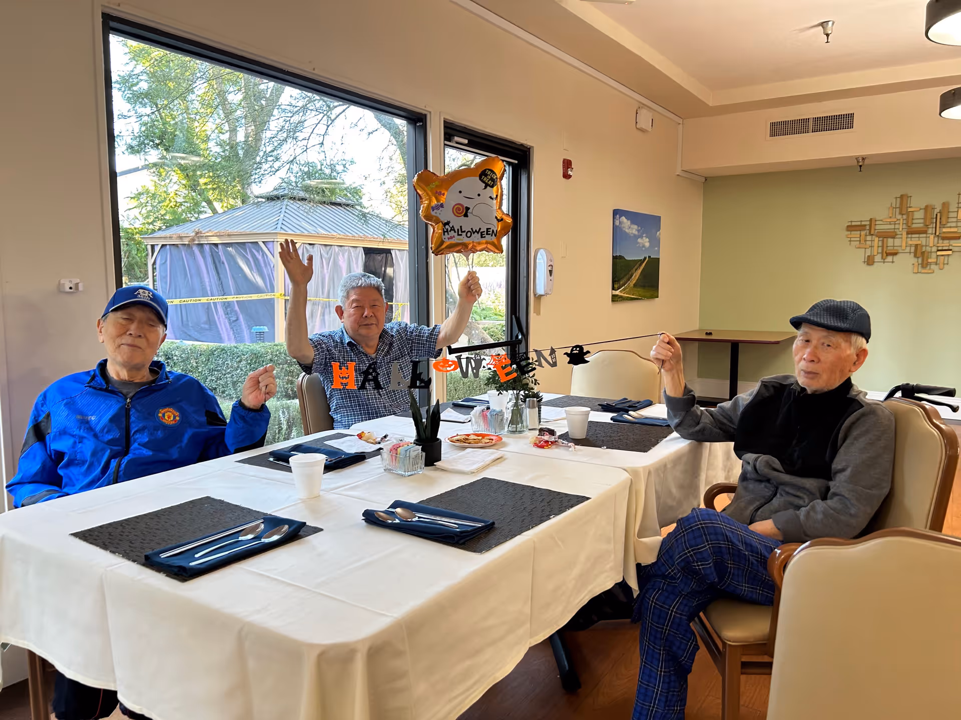 Three elderly men sitting around a dining table decorated with a Halloween banner and a Halloween-themed balloon. The table is set with placemats, utensils, and cups. Large windows behind them show greenery and a gazebo outside.