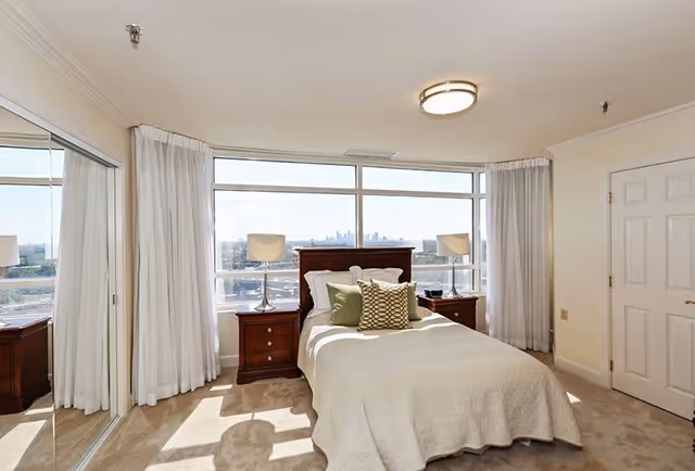 A bright bedroom with a large window offering a city view. The room features a bed with white bedding and decorative pillows, two wooden nightstands with matching lamps, white curtains, a mirrored closet door, and a closed white door.