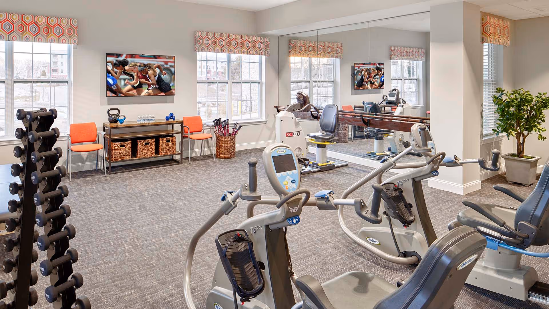 Well-lit fitness room with exercise machines, a rack of dumbbells, mirrored wall, TV, seating and windows.