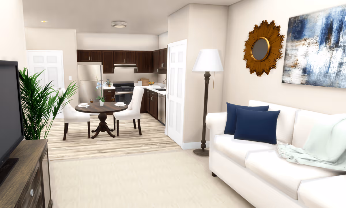 A bright and modern living area featuring a white sofa with blue pillows and a light throw, a tall floor lamp, a decorative sunburst mirror, and abstract wall art. Adjacent to the living area is a small dining space with a round wooden table set for two and two white upholstered chairs. The kitchen in the background has dark wood cabinets, stainless steel appliances, and light wood flooring.
