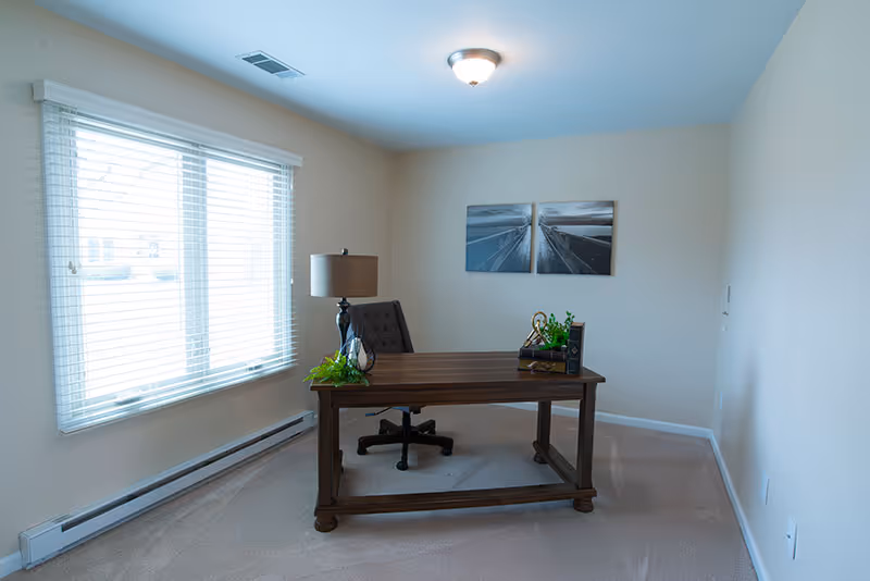 A small office room with beige walls and carpeted floor. There is a wooden desk with a black office chair behind it. On the desk are a table lamp, a small plant, and some decorative items. A large window with white blinds is on the left wall, and two black and white framed pictures hang on the wall behind the desk.