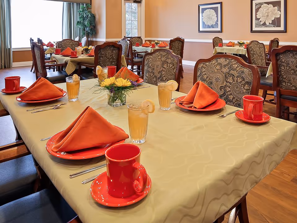 A dining room set up with tables covered in beige tablecloths, each table set with red plates, red cups and saucers, orange folded napkins, glasses of iced tea with lemon, and small floral centerpieces. The room has patterned upholstered chairs, wooden flooring, and framed floral artwork on the walls.