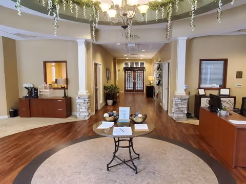 Spacious senior living lobby with a central round table, reception desks, decorative columns, hanging greenery, and a hallway leading to double doors.