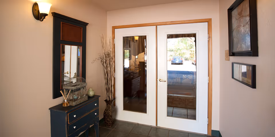 A small entryway area with double glass doors framed in wood. To the left is a dark wooden dresser with a mirror above it, decorated with a birdcage, a small ceramic bird, and reed diffuser sticks. A tall vase with decorative branches stands in the corner. On the right wall, there are two framed pictures. The floor is tiled and a wall sconce light fixture is mounted above the dresser.