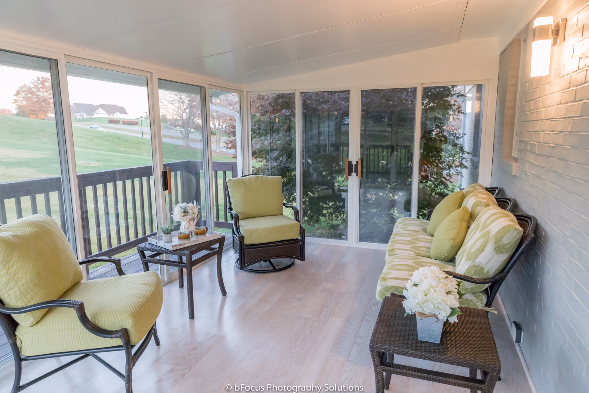 A bright sunroom with large sliding glass doors and windows offering a view of a green lawn and trees outside. The room features light wood flooring, a cushioned sofa with green and patterned pillows, two cushioned armchairs, and two small wicker tables with decorative plants and flowers. A wall-mounted light fixture is visible on the right side.