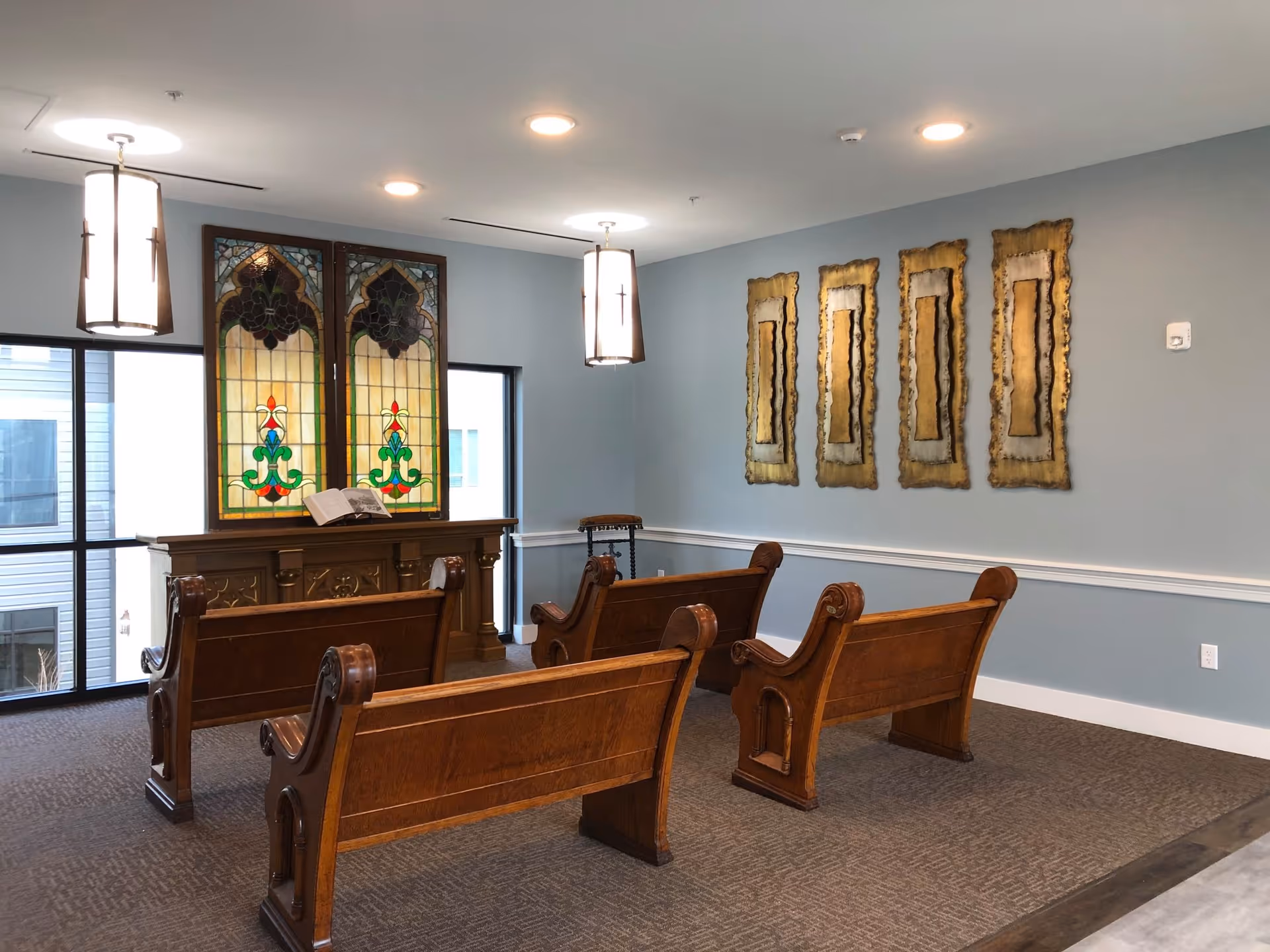 Interior room with wooden pews arranged facing a wooden altar with stained glass panels behind it. The room has light blue walls with four decorative gold-framed wall hangings and two hanging pendant lights from the ceiling.