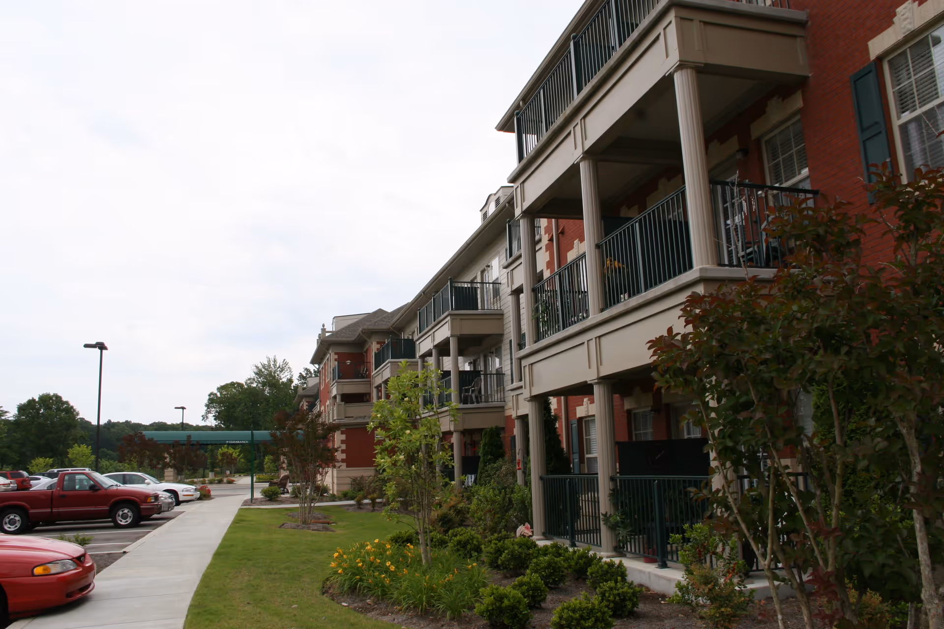Exterior view of a multi-story senior living community building with balconies, landscaped greenery, and a parking lot with several cars parked along a sidewalk.