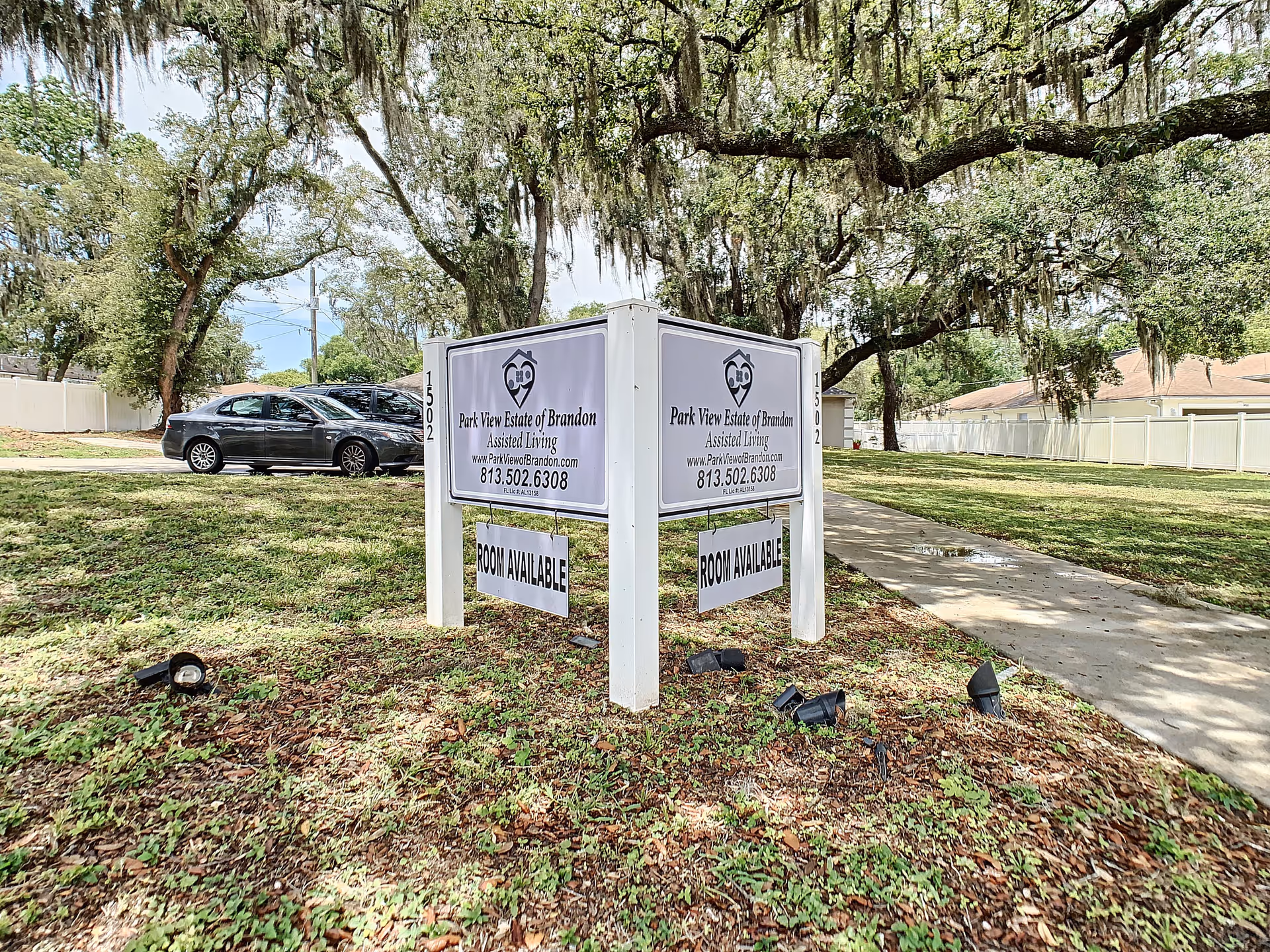 A freestanding sign reading 'Park View Estate of Brandon Assisted Living' on a grassy lot beneath large moss-draped oak trees, with parked cars and a sidewalk in the background.