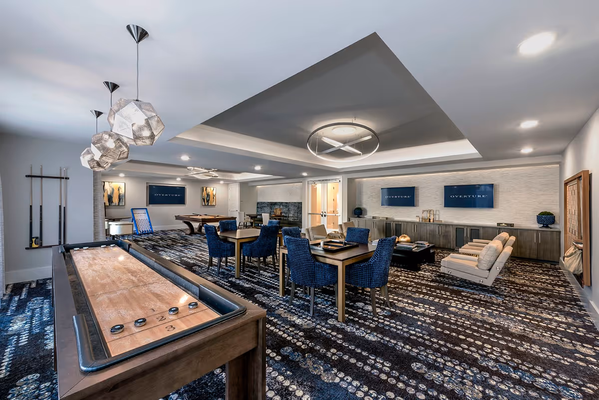 A spacious and modern game room with a shuffleboard table in the foreground, several tables with blue upholstered chairs, a pool table in the background, and a seating area with beige armchairs. The room features contemporary lighting fixtures, patterned carpet, and multiple screens displaying the word 'OVERTURE'.