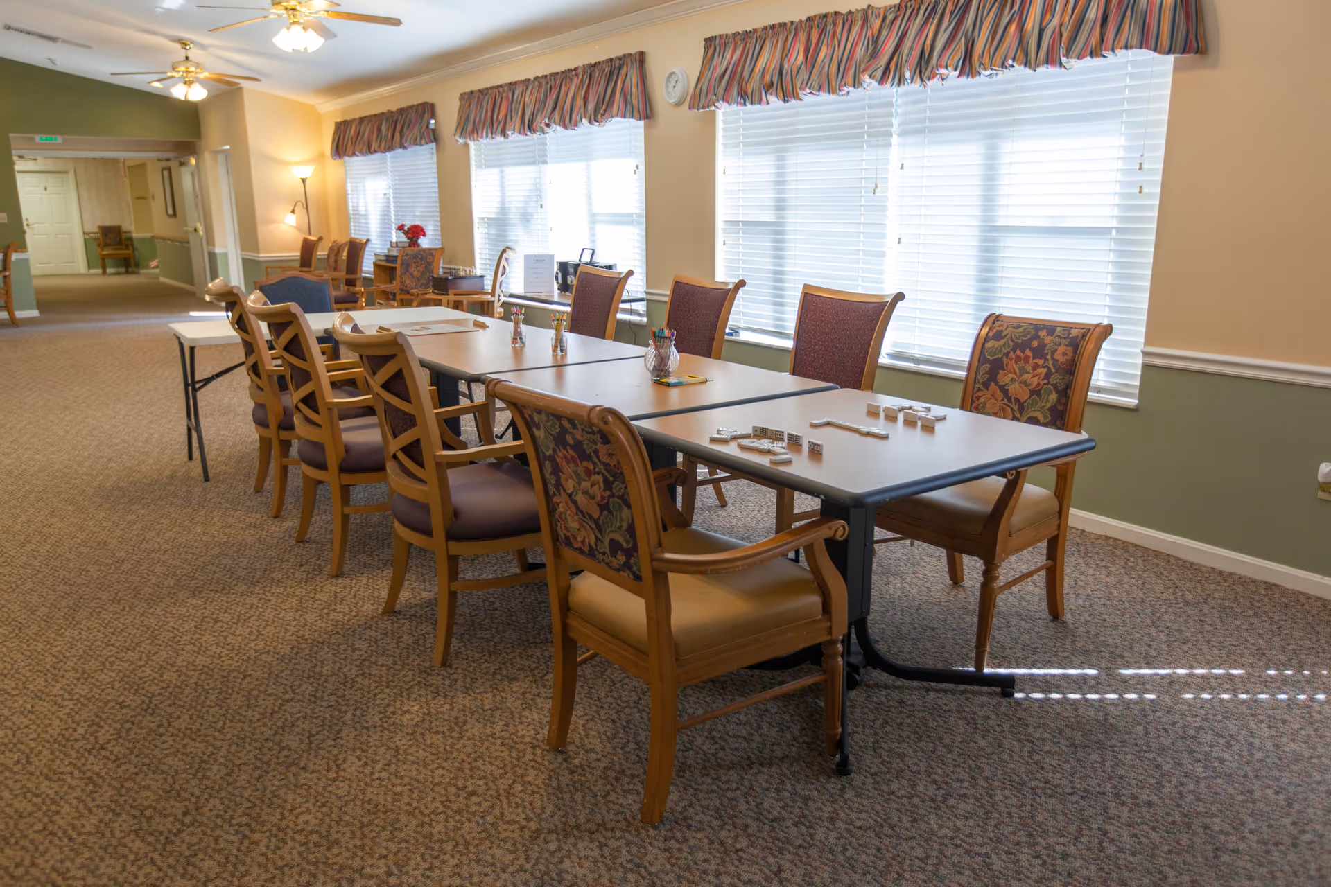 A well-lit room with a long table surrounded by wooden chairs with cushioned seats and backs. The table has dominoes and small containers with colored pencils on it. Large windows with blinds and striped valances let in natural light. The room has carpeted floors and light-colored walls with a green accent wall in the background.