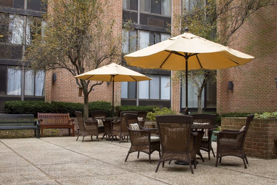 Outdoor patio area with multiple wicker chairs and tables under large beige umbrellas, surrounded by brick building walls, trees, and benches.