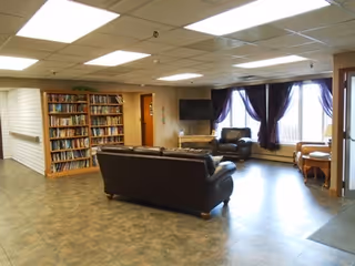 A spacious living room area with two dark leather sofas, a flat-screen TV mounted on the wall, a bookshelf filled with books, large windows with dark curtains, and a small table with chairs near the windows.