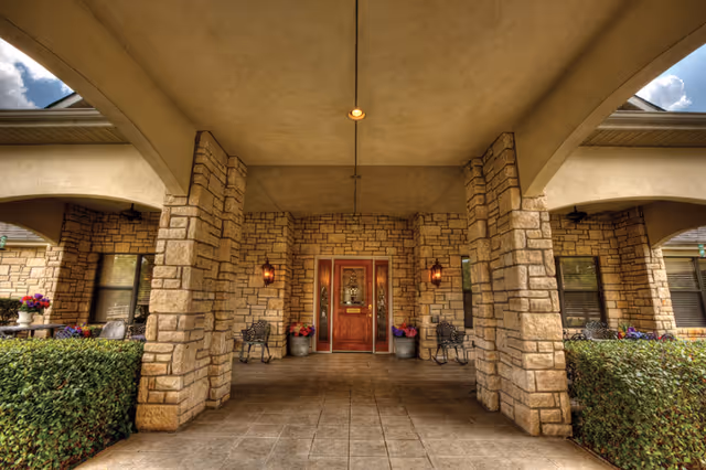 Covered entrance area of a building with stone pillars and walls, a wooden door in the center flanked by two lantern-style wall lights, metal chairs on either side, and potted flowers near the door and along the sides.