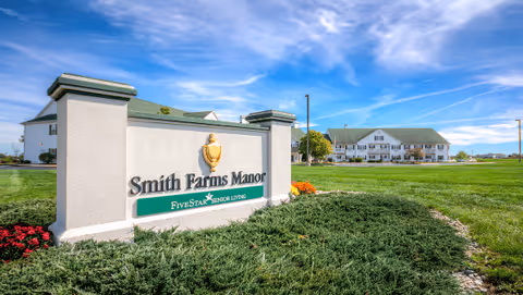 Entrance sign reading 'Smith Farms Manor' in front of a landscaped lawn and a large white multi-story senior living building under a blue sky.