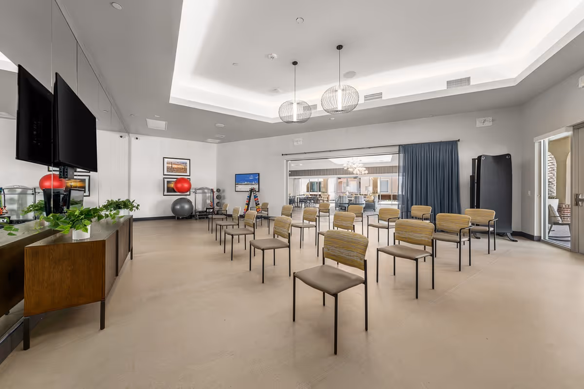 Large, well-lit community activity room with rows of chairs, a wall-mounted TV, and exercise equipment.