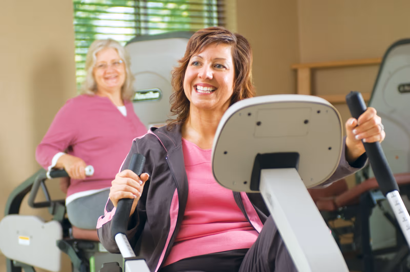 Two women exercising on fitness machines in a well-lit indoor gym or exercise room. The woman in the foreground is smiling and using a rowing machine, while the woman in the background is also engaged in exercise equipment.