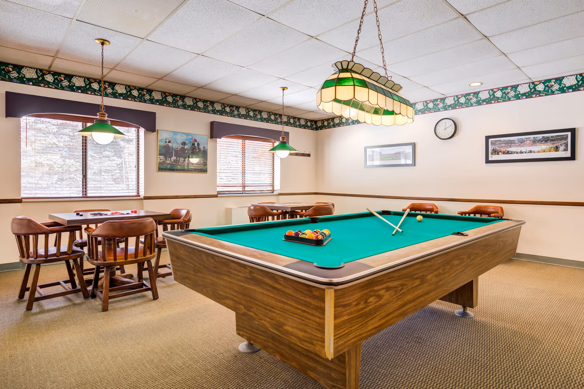 A recreational room with a pool table in the center, two pool cues resting on the table, and a rack of billiard balls. The room has carpeted flooring, two windows with blinds, and several wooden chairs around small tables. The walls are decorated with framed pictures and a clock, and there are hanging lights above the pool table and tables.