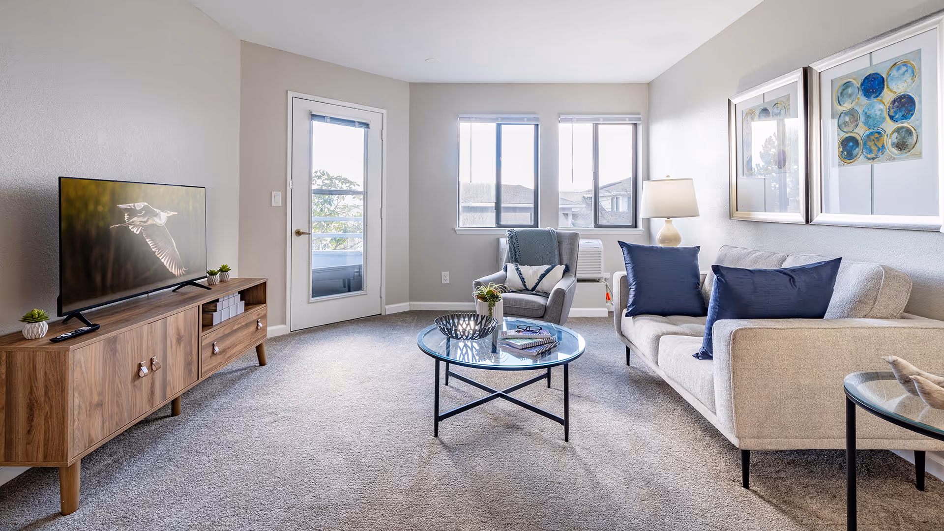 A bright and modern living room with a beige sofa adorned with blue pillows, a gray armchair with a throw blanket, a round glass coffee table with books and decor, a wooden TV stand with a flat-screen TV displaying a bird in flight, and two framed artworks on the wall. The room has carpeted flooring, three windows, and a glass door leading outside.