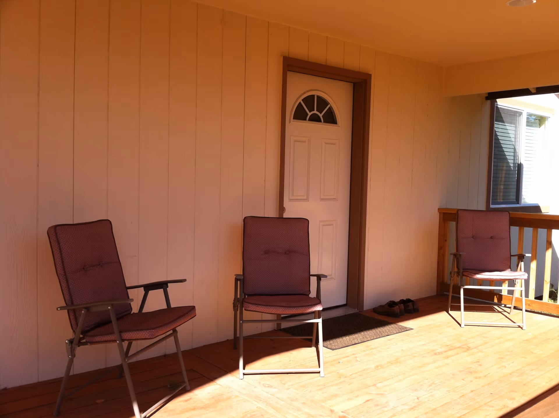 A covered wooden porch with three cushioned chairs arranged in front of a white door with a small arched window. A pair of shoes is placed on a mat near the door, and there is a window to the right side of the porch.