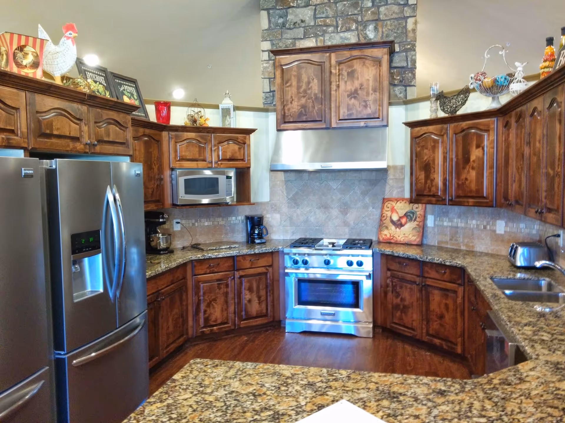 A spacious kitchen with dark wooden cabinets, granite countertops, and stainless steel appliances including a refrigerator, microwave, and stove. The stove is set against a stone backsplash with a stainless steel hood above it. Various decorative items such as a rooster figurine and framed pictures are placed on top of the cabinets.