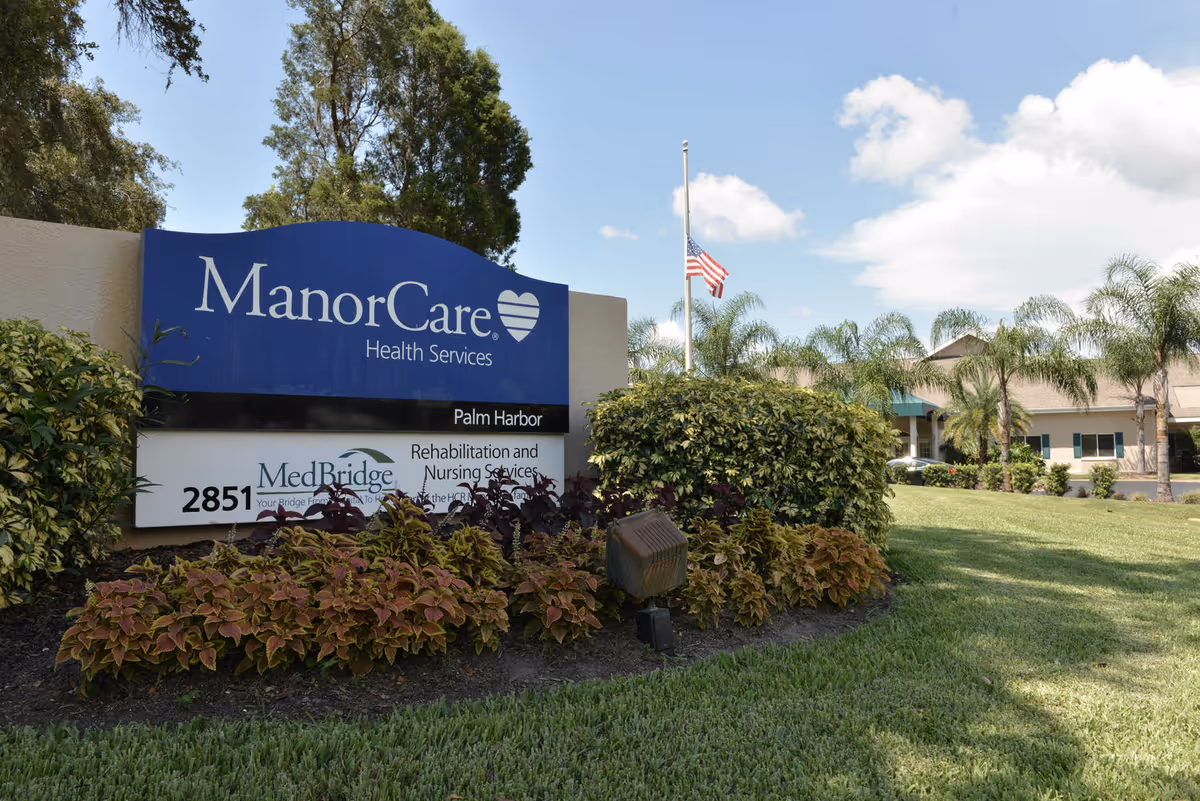 Outdoor view of a health services facility sign for ManorCare Health Services in Palm Harbor, surrounded by bushes and plants with a building and palm trees in the background under a partly cloudy sky.