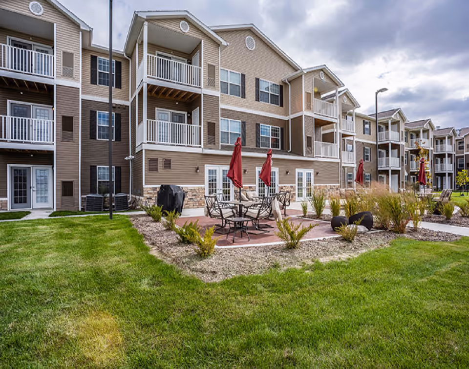 Exterior view of a multi-story residential building with balconies and patios. The foreground features a well-maintained lawn, landscaped garden beds, and outdoor seating areas with tables and red umbrellas.