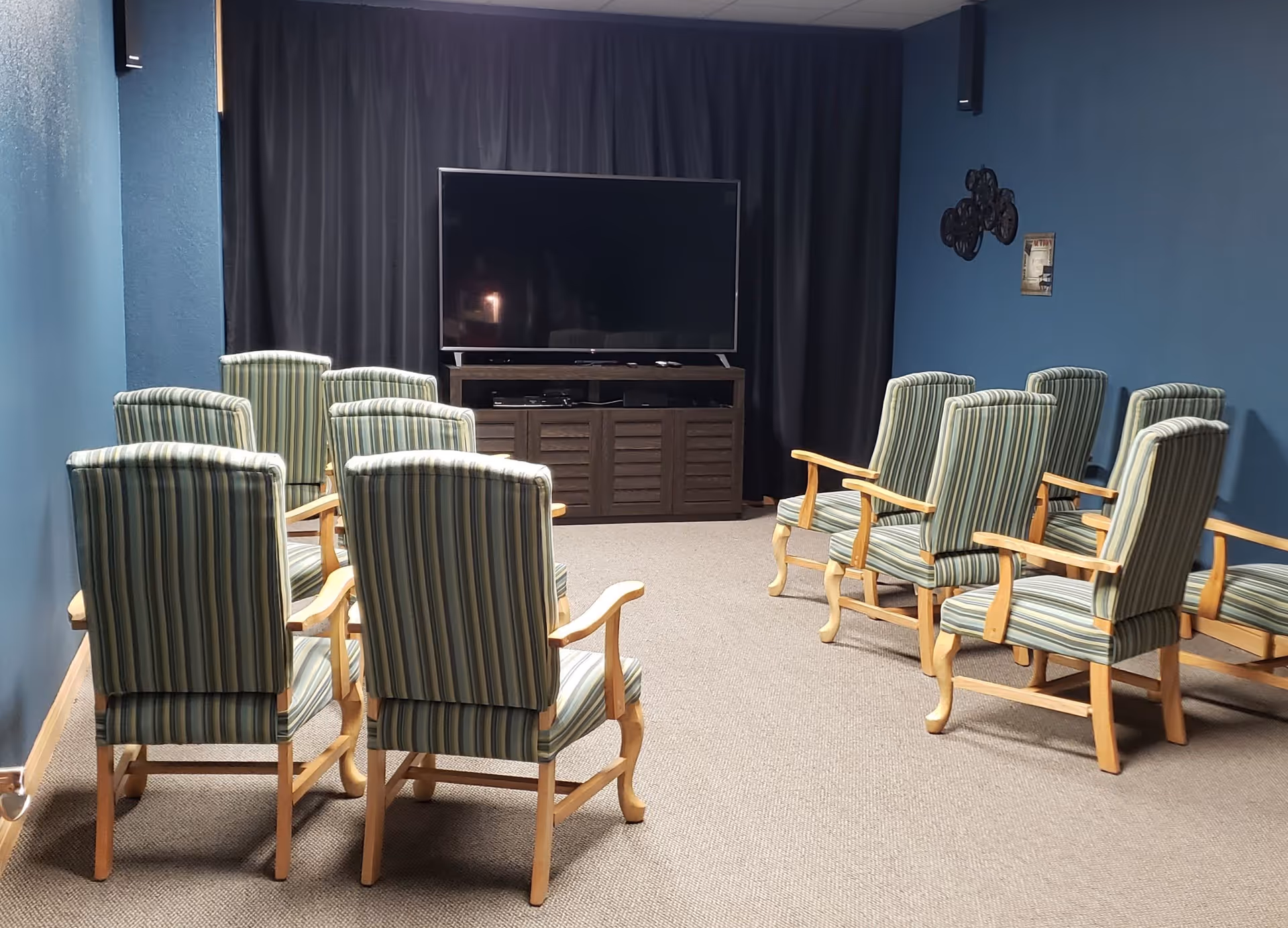 A small media room with two rows of striped armchairs facing a large flat-screen TV mounted on a dark wooden cabinet. The walls are painted blue, and there is a black curtain behind the TV. The carpet is beige, and there are a few wall decorations on the right side.