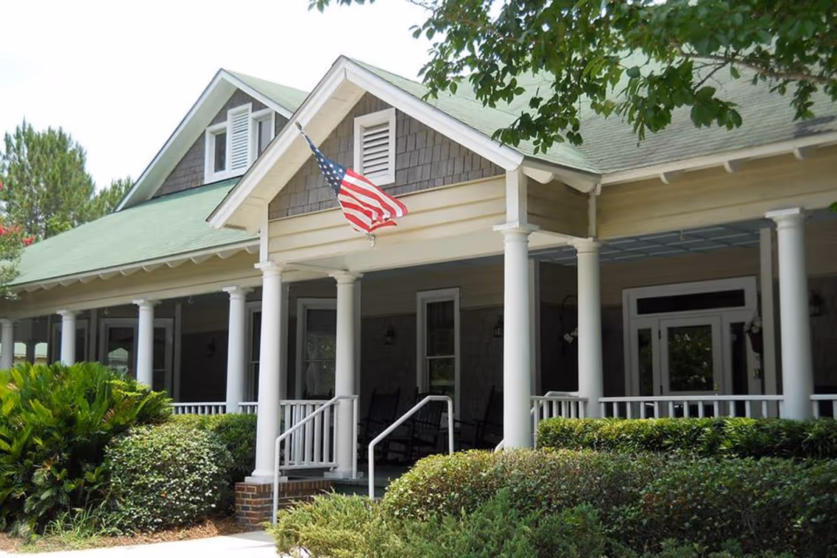 Front exterior view of a building with a green roof, white columns, and a porch with rocking chairs. An American flag is mounted on the front of the building. There are bushes and trees surrounding the entrance.