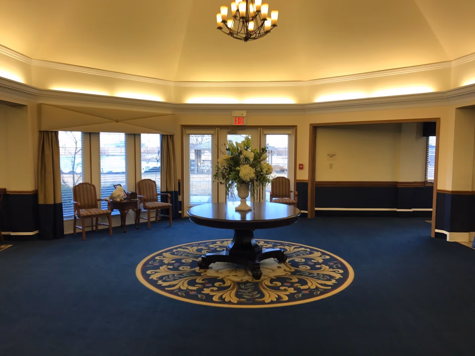 A spacious interior room with a round wooden table in the center holding a large floral arrangement. The floor is carpeted with a blue and gold patterned rug under the table. There are several wooden chairs with striped cushions arranged along the walls near large windows and glass doors. The ceiling is high with soft lighting and a chandelier hanging above the table.