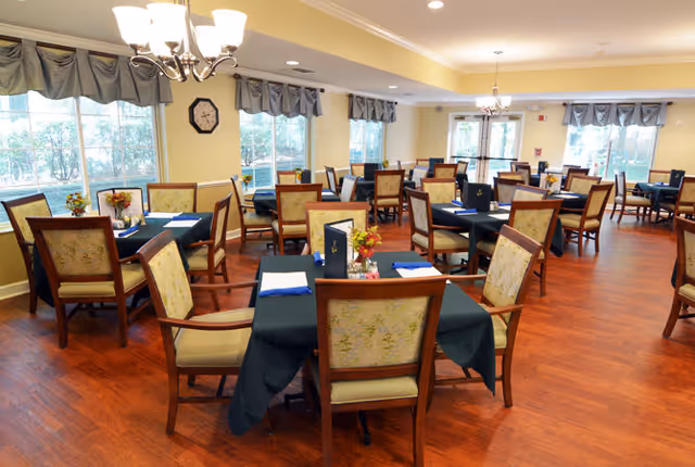 Bright dining room with multiple set tables, upholstered chairs, chandeliers, and large windows.