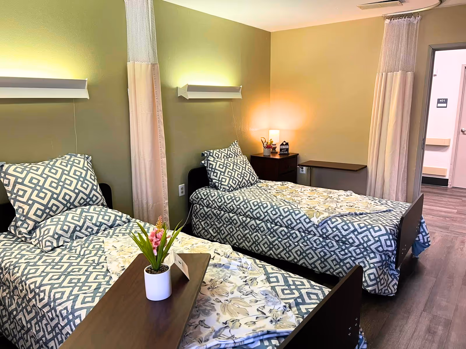 A nursing facility room with two single beds, each with patterned bedding and pillows. There are privacy curtains between the beds and a small nightstand with a lamp and decorative items. The room has light green walls and wood flooring, with a door leading to a hallway.