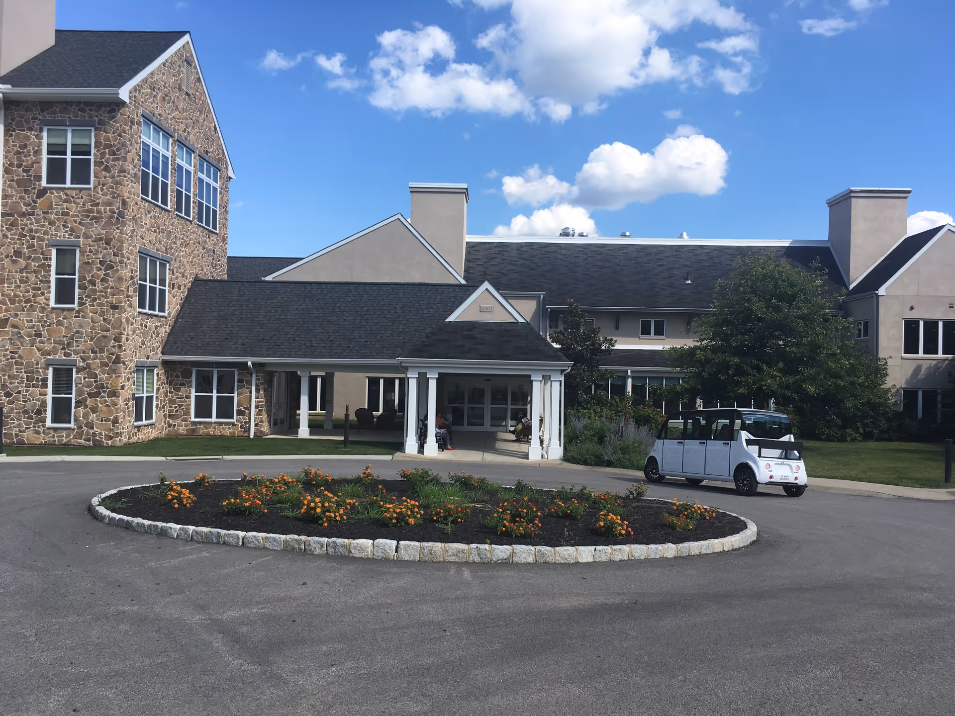 Exterior view of a senior living facility with a stone and beige building, a covered entrance, a circular flower bed with orange flowers in front, and a small white electric shuttle vehicle parked on the driveway under a blue sky with scattered clouds.