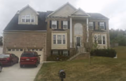 Front exterior view of a large two-story brick house with a double garage, multiple windows, a central entrance with steps, and a sloped lawn with a mailbox in front.
