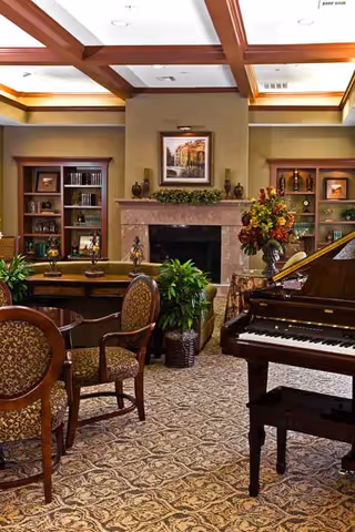 Elegant senior living lounge with fireplace, piano, bookshelves, plants, and patterned carpet.