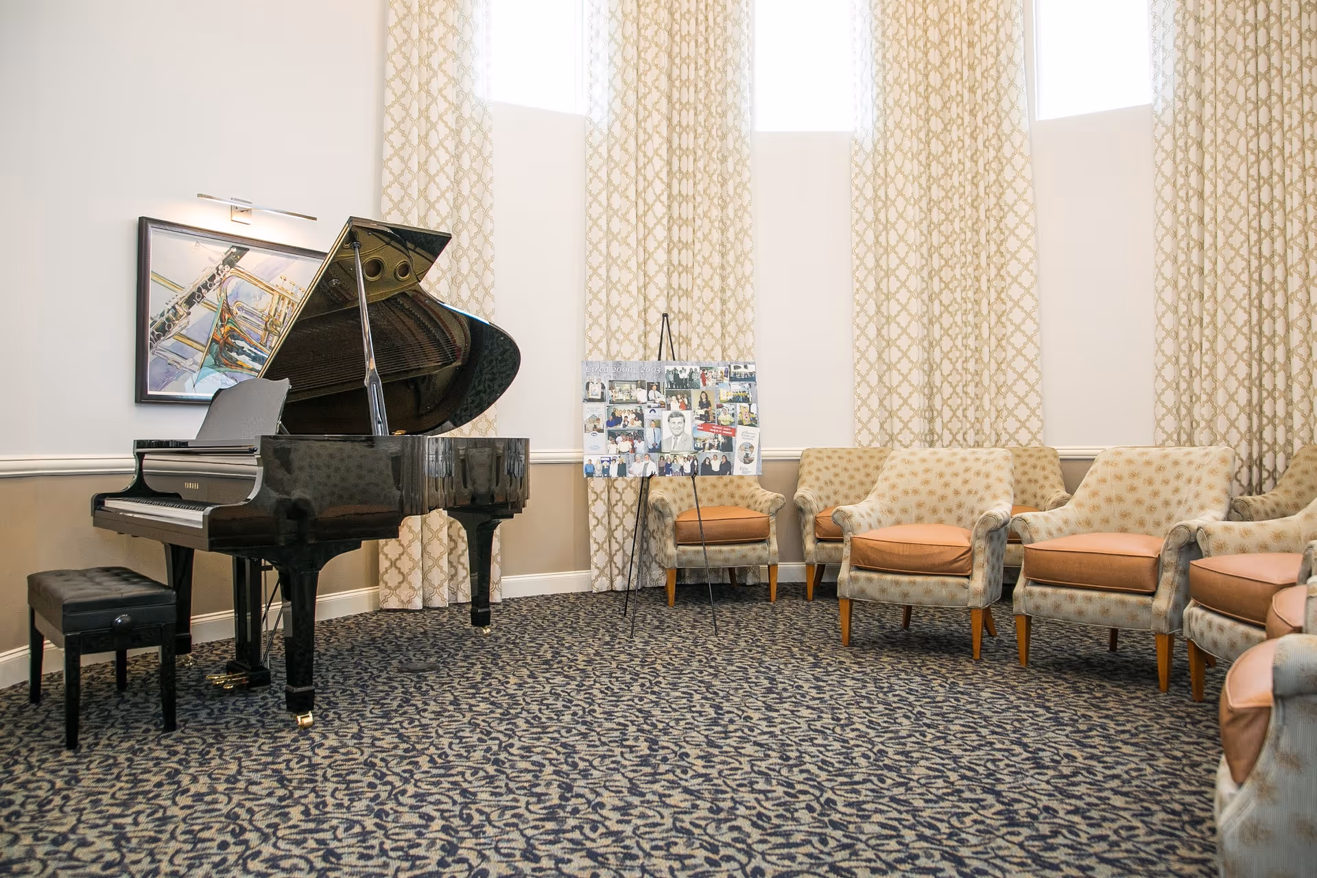 A bright common room with a black grand piano on the left and a row of upholstered chairs against patterned curtains.
