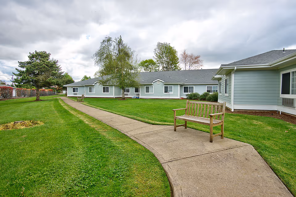 Outdoor view of a senior living facility with a concrete pathway leading through a well-maintained green lawn. There are wooden benches along the path and several trees scattered around. The building is light blue with white trim and multiple windows. The sky is cloudy.