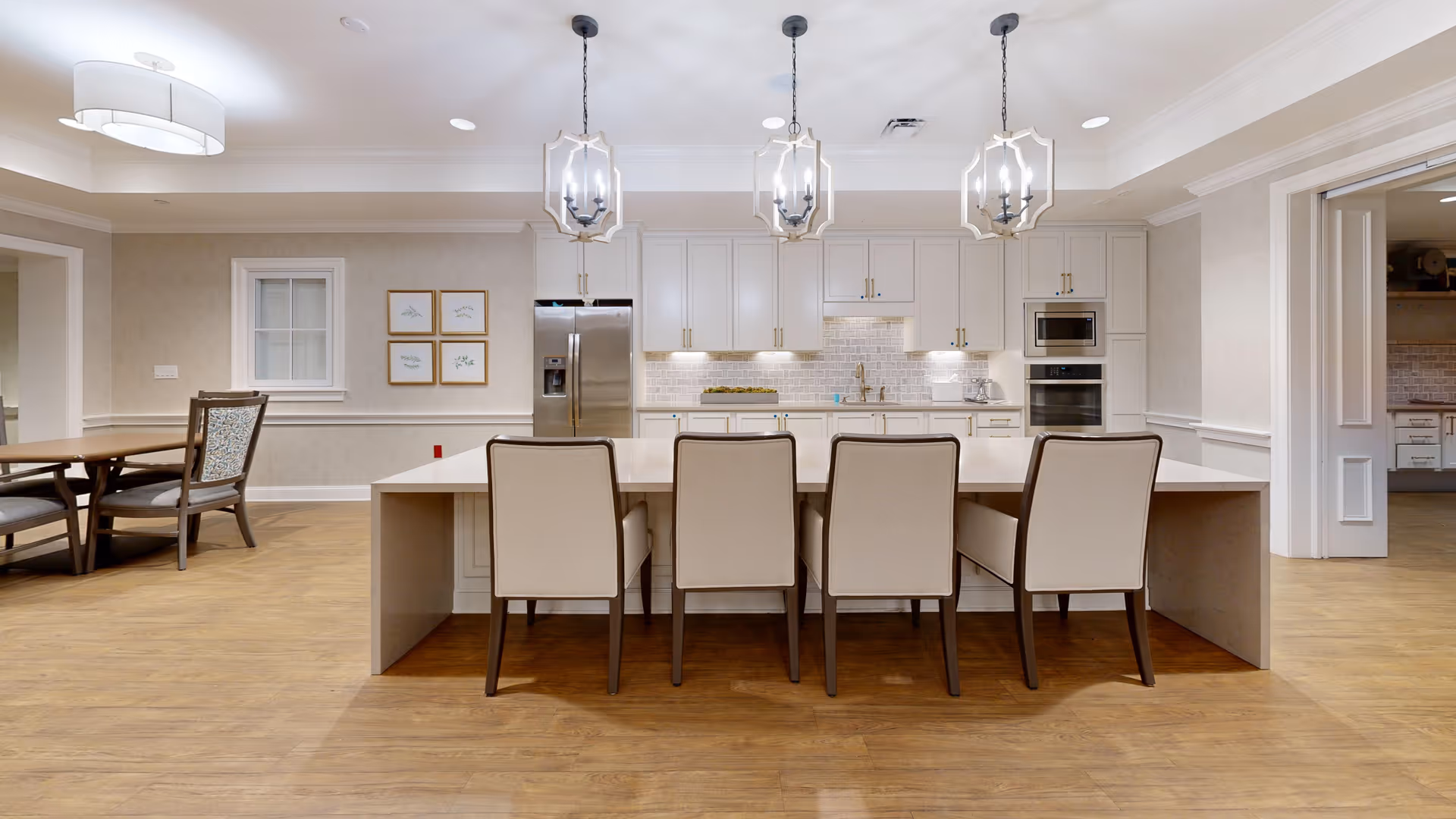 A modern kitchen with a large island featuring four beige chairs. The kitchen has white cabinetry, a stainless steel refrigerator, built-in microwave and oven, and three pendant lights hanging above the island. To the left, there is a dining area with a wooden table and chairs. The floor is wood, and the walls are light-colored with framed artwork.