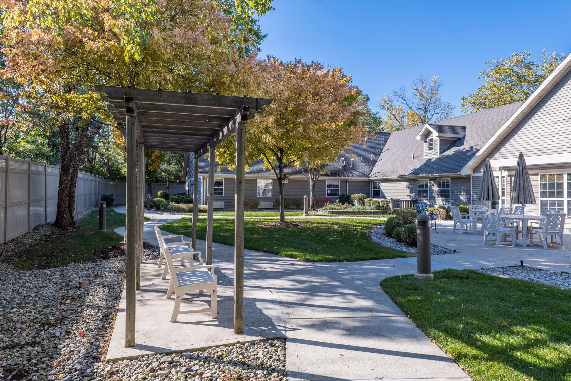 Outdoor courtyard area at Arden Courts - ProMedica Memory Care Community (Bingham Farms) featuring a wooden pergola with benches underneath, a paved walkway, green grass, trees with autumn foliage, and a patio area with tables, chairs, and umbrellas beside a gray building under a clear blue sky.