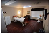 A simple, clean room with a hospital-style bed, a white dresser, a brown armchair, a small round table with a lamp, a wall-mounted TV, and a window with blinds and a dark valance. The floor is dark wood, and the walls are light-colored.