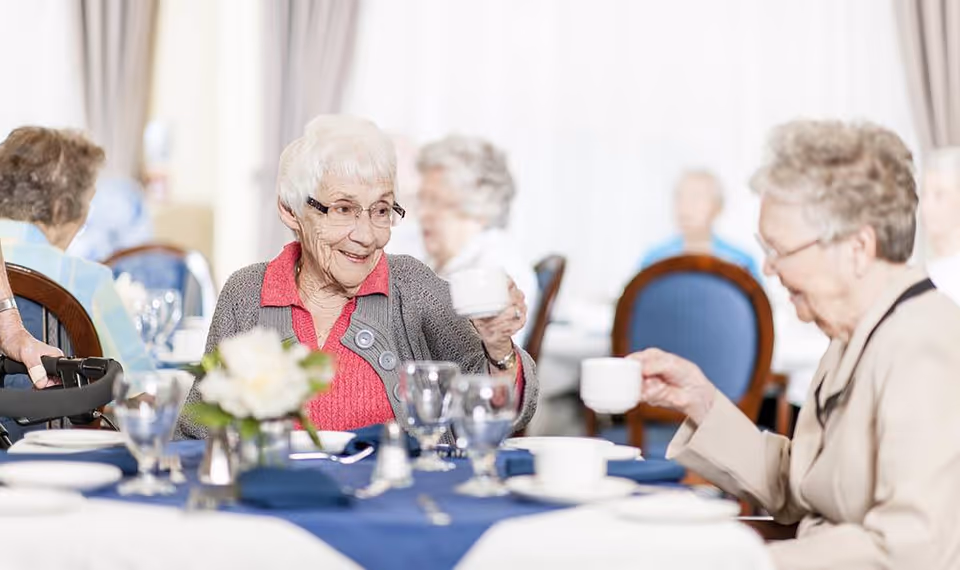 Two elderly women sitting at a dining table in a senior living facility, smiling and holding up cups as if toasting. The table is set with glassware, plates, and a flower centerpiece. Other elderly individuals are visible in the background seated at tables.