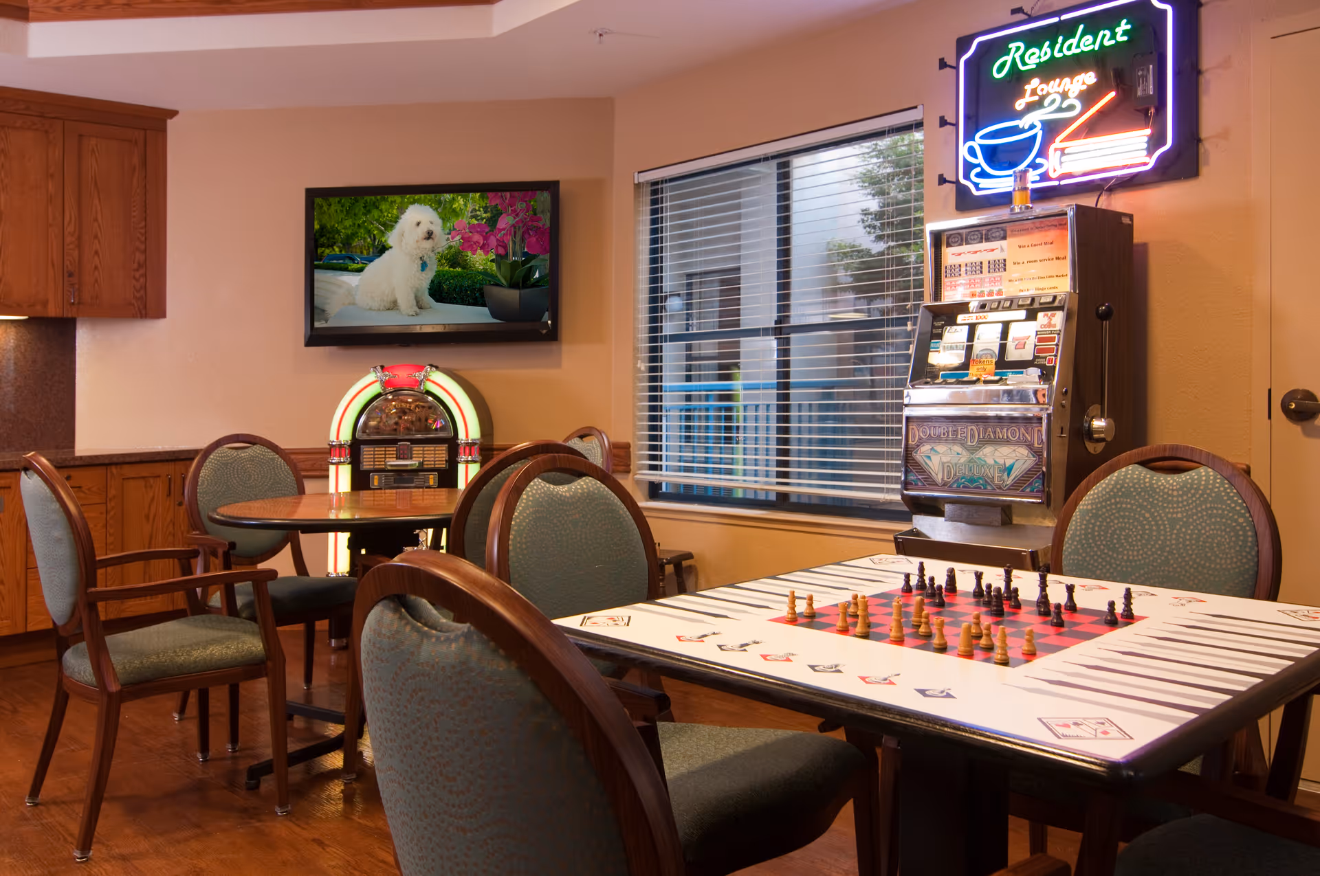 A senior living facility game room with tables and chairs, a chessboard on a table, a jukebox, a slot machine and a wall-mounted TV.