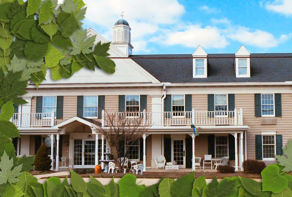 Exterior view of a two-story senior living facility building with beige siding, white trim, and green shutters. The building features a covered porch with white chairs and tables, a small balcony on the second floor, and a cupola on the roof. Green leaves frame the top left and bottom edges of the image, with a blue sky and clouds in the background.