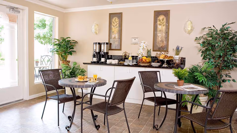 A cozy dining area with two round tables and four wicker chairs. Each table has a small potted plant, and one table has a newspaper. In the background, there is a counter with coffee dispensers, cups, a basket of fruit, and pastries. The room is decorated with framed botanical artwork and several green plants. A glass door and window let in natural light from outside.