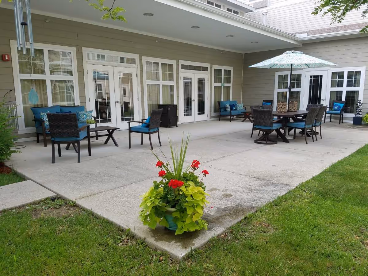 Outdoor patio area with multiple seating arrangements including chairs with blue cushions, a table with an umbrella, and a planter with red flowers and green foliage in the foreground. The patio is adjacent to a building with multiple glass doors and windows.
