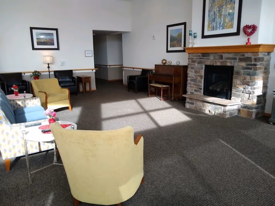 A cozy senior living common area with several armchairs arranged around small tables, a stone fireplace with a wooden mantle decorated with framed artwork and a heart-shaped ornament, and a piano with a bench. The room is carpeted and well-lit by natural sunlight coming through windows.