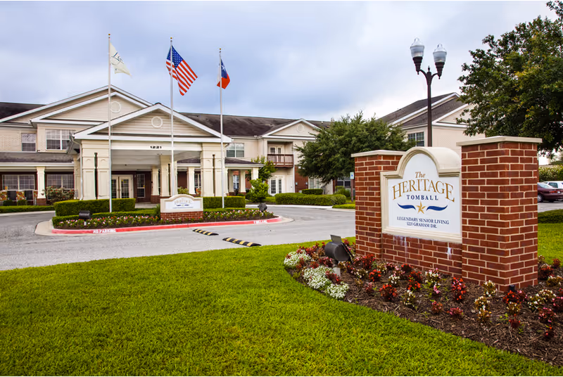 Exterior view of The Heritage Tomball Senior Living facility showing the main entrance with three flagpoles displaying flags, a well-maintained lawn, flower beds, and a brick sign with the facility's name and address.