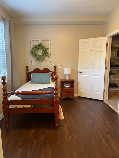 A cozy bedroom with a wooden single bed featuring a white mattress, a blue pillow, and a gray blanket. Next to the bed is a wooden nightstand with a white lamp and some decorative items. A window with light gray curtains is on the left wall, and a decorative wreath hangs above the bed. An open door reveals a closet with shelves on the right side. The room has wooden flooring and beige walls.