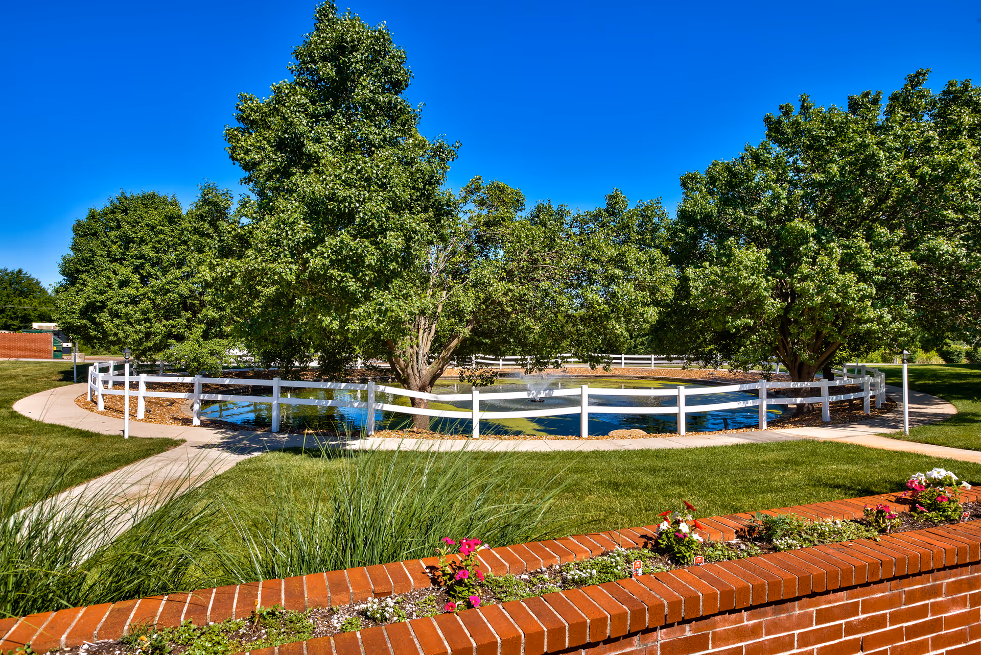 A landscaped outdoor area with a circular pond surrounded by a white fence, mature trees, a brick flower bed and paved walkways under a clear blue sky.