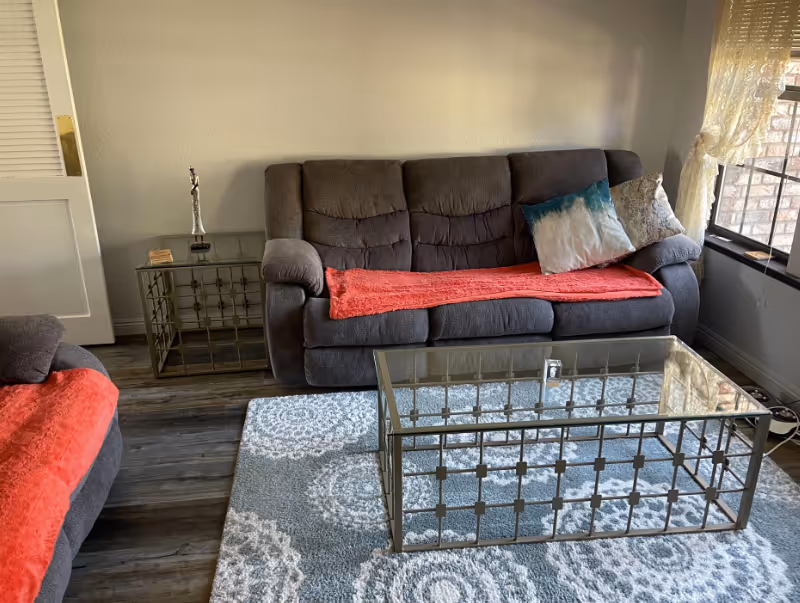 Cozy living room with a gray upholstered sofa and matching chair, glass coffee table, patterned rug, and a window with lace curtains.