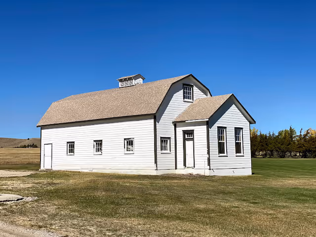 A white wooden building with a gabled roof and several windows, situated on a grassy area under a clear blue sky. The building has a small entrance porch and appears to be in a rural or open landscape setting.